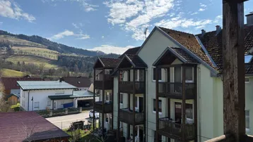 Blick von einem Balkon auf ein Wohngebäude mit weiteren Balkonen und bewaldeten Hügeln im Hintergrund.