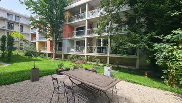 Außenbereich mit Kiesterrasse und Gartenmöbeln vor einem modernen Wohngebäude mit Balkonen.