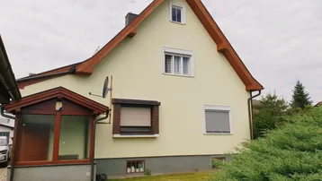 Hausfassade mit hellgelbem Putz, braunem Dach und kleinen Fenstern in einer ruhigen Wohngegend.