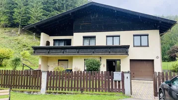 Traditionelles Haus mit Holzbalkon und Garage, umgeben von grünen Bäumen und Natur.
