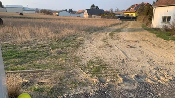 Weitläufiges unbebautes Feld mit einem Feldweg und Häusern in der Ferne unter blauem Himmel.