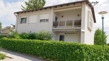 Hausfassade mit einem Balkon im Obergeschoss, umgeben von einer grünen Hecke.