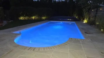 Beleuchteter Swimmingpool bei Nacht, umgeben von einer gepflasterten Terrasse und üppigem Grün.