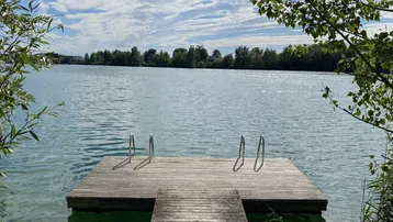Holzsteg mit Badeleiter am Seeufer, umgeben von grünen Bäumen unter blauem Himmel.