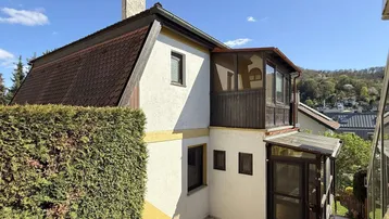 Seitenansicht des Hauses mit Wintergarten, Balkon und gepflegter Hecke unter blauem Himmel.