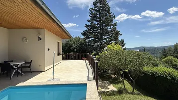Große Terrasse mit Pool und Sitzbereich, umgeben von grüner Natur unter blauem Himmel.