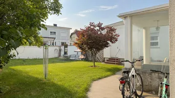 Grüner Gartenbereich mit Rasenfläche, Bäumen und Sitzgelegenheiten im Hintergrund.