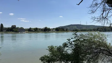 Weitläufiger Blick über einen Fluss mit bewaldeten Hügeln im Hintergrund unter blauem Himmel.