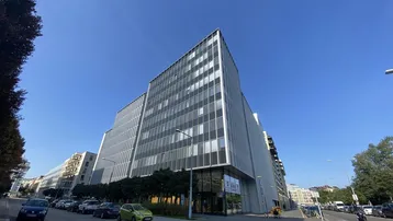 Modernes Bürogebäude mit Glasfassade und klarem Himmel, umgeben von einer städtischen Straße mit parkenden Autos.