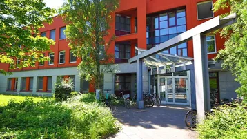 Eingangsbereich eines modernen Bürogebäudes mit viel Grün und Fahrradstellplätzen unter einem Vordach.