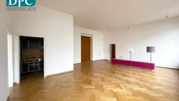 Geräumiger, heller Raum mit Fischgrätparkett und Zugang zu einer kleinen Küche.