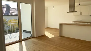 Heller offener Wohn-Essbereich mit moderner Einbauküche, Holzboden und Zugang zu einem Balkon.