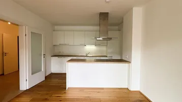 Heller offener Wohnbereich mit moderner Einbauküche, Holzboden und Zugang zu weiteren Räumen.