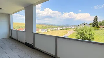 Geräumiger Balkon mit Fliesenboden und weitem Blick über die grüne Landschaft und umliegende Häuser.