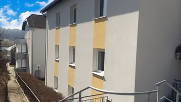 Modernes Mehrfamilienhaus mit heller Fassade, gelben Akzenten und Zugang über eine Rampe.
