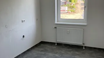 Leeres Zimmer mit grauen Fliesen, einem Fenster und Heizkörper, bereit für die individuelle Gestaltung.