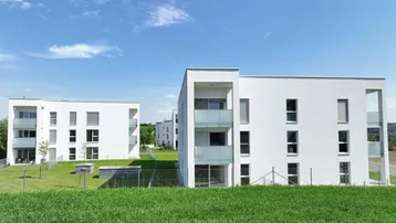 Moderne weiße Wohngebäude mit Balkonen, umgeben von grünen Wiesen unter blauem Himmel.