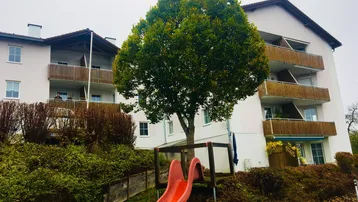 Mehrfamilienhaus mit Balkonen und einem Spielplatz im Vordergrund, umgeben von Bäumen und Büschen.