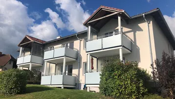 Mehrfamilienhaus mit mehreren Balkonen und gepflegter Grünfläche unter blauem Himmel.