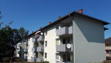 Mehrfamilienhaus mit mehreren Balkonen unter blauem Himmel und grüner Umgebung.