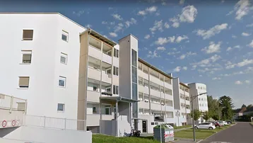 Modernes Mehrfamilienhaus mit weißer Fassade, Balkonen und umliegenden Grünflächen unter blauem Himmel.