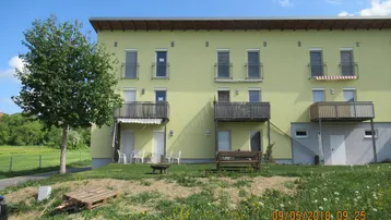 Gelbes Mehrfamilienhaus mit mehreren Balkonen und einer Terrasse, umgeben von einer grünen Wiese.