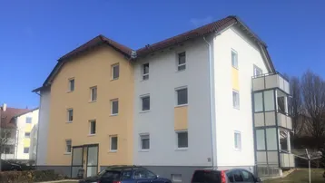 Mehrfamilienhaus mit heller Fassade, Satteldach und Balkonen unter blauem Himmel.