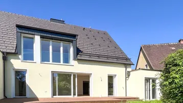Moderne Hausrückseite mit großer Holzterrasse und Zugang zum Garten unter blauem Himmel.