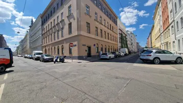 Blick auf eine belebte Straßenecke in der Stadt mit traditionellen Mehrfamilienhäusern unter blauem Himmel.