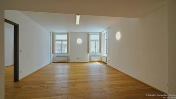 Heller, leerer Raum mit Holzboden, weißen Wänden und zwei Fenstern, die viel Tageslicht hereinlassen.