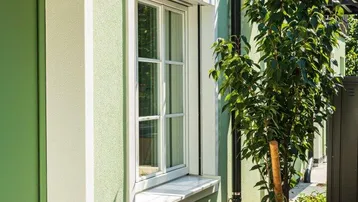 Grüne Fassade eines Gebäudes mit Fenster, kleinem Gartenbereich und Holztisch im Freien.
