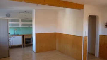 Offener Wohnbereich mit Holzboden, sichtbaren Holzbalken und angrenzender Küche mit hellen Schränken.