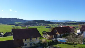 Weitläufige Landschaftsaufnahme mit mehreren Häusern, grünen Feldern und einer Bergkulisse im Hintergrund.