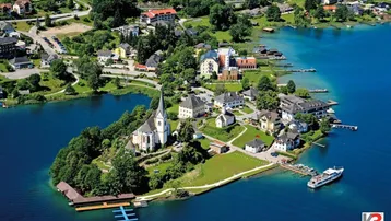 Luftaufnahme eines malerischen Dorfes auf einer Halbinsel am See mit umliegender grüner Landschaft und Bergen.