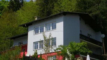 Mehrstöckiges Wohnhaus mit grauer und roter Fassade, umgeben von üppiger grüner Natur und Bäumen.