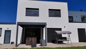 Moderne Hausfassade mit überdachter Terrasse, Rasenfläche und Gartenmöbeln.