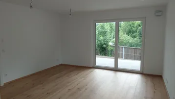 Helles Zimmer mit Holzboden und großem Fenster, das Zugang zu einer Terrasse bietet und ins Grüne blickt.