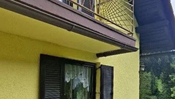 Gelbe Hausfassade mit braunem Sockel, einem Balkon mit Ziergeländer und Fensterläden.