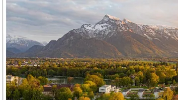 Panoramablick auf schneebedeckte Berge, einen See und eine grüne Landschaft mit modernen Gebäuden.