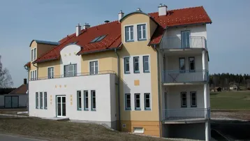 Modernes Mehrfamilienhaus mit gelb-weißer Fassade, roten Ziegeldächern und mehreren Balkonen, umgeben von grüner Landschaft.