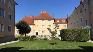Mehrfamilienhaus mit rotem Ziegeldach, umgeben von einem gepflegten Rasen und hohen Hecken in einem sonnigen Innenhof.