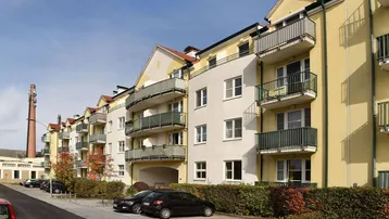 Mehrfamilienhaus mit gelber und weißer Fassade, Balkonen und Parkplätzen vor dem Gebäude.