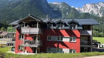 Modernes Mehrfamilienhaus mit roter Fassade, Balkonen und beeindruckendem Bergblick in alpiner Umgebung.