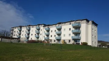 Mehrfamilienhaus mit mehreren Balkonen und weitem Blick über grüne Wiesen unter blauem Himmel.