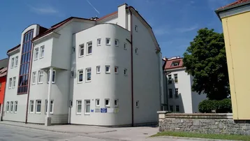 Modernes Gebäude mit heller Fassade, vielen Fenstern und rotem Ziegeldach an einer Straßenecke.