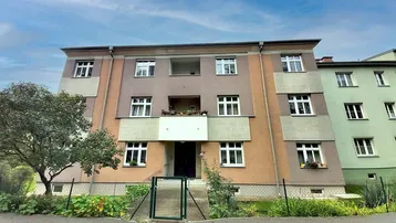 Mehrfamilienhaus mit mehreren Etagen und Balkonen, umgeben von Grünflächen unter blauem Himmel.