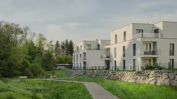 Moderne weiße Wohngebäude mit Balkonen, umgeben von Grünflächen und Bäumen in ländlicher Umgebung.