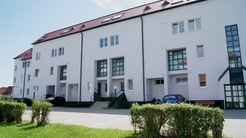 Mehrfamilienhaus mit weißer Fassade, rotem Mansarddach und Stellplätzen im Vorgartenbereich.