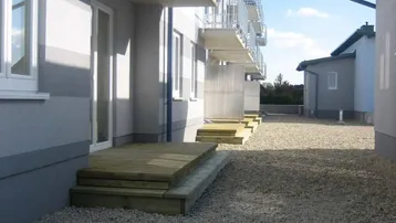 Außenansicht eines Mehrfamilienhauses mit mehreren Balkonen und ebenerdigen Holzterrassen auf Kiesfläche.