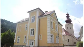 Historisches Gebäude mit gelber Fassade und weißer Stuckverzierung, neben einem Kirchturm gelegen.